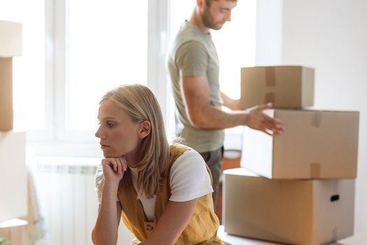 Eine Frau sitzt nachdenklich zwischen Umzugskisten, die ihr Ex-Mann packt